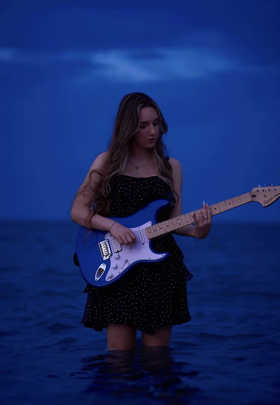 Liza holding a blue electric guitar while standing in the water at dusk